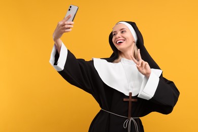 Photo of Happy woman in nun habit taking selfie and showing peace sign against orange background