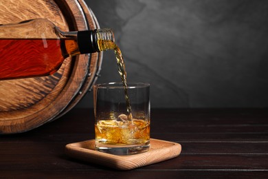 Pouring whiskey from bottle into glass with ice cubes near wooden barrel on table, space for text