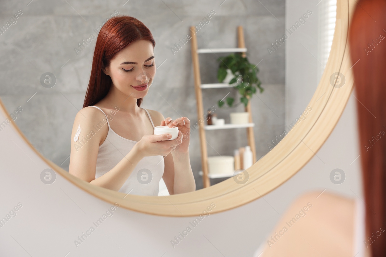 Photo of Beautiful young woman applying body cream onto shoulder near mirror indoors