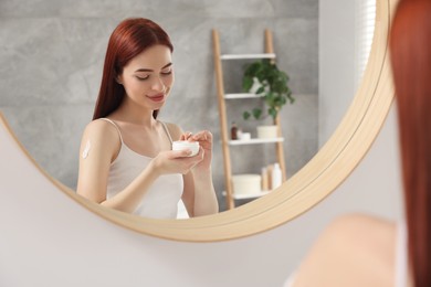 Photo of Beautiful young woman applying body cream onto shoulder near mirror indoors