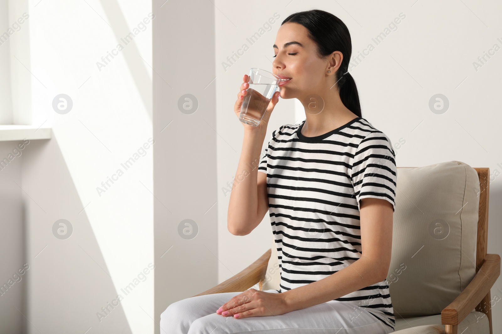 Photo of Young woman drinking water indoors, space for text. Refreshing drink