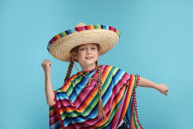 Photo of Cute girl in Mexican sombrero hat and poncho dancing on light blue background