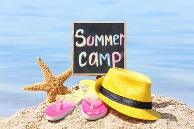 Photo of Little blackboard with text SUMMER CAMP, hat and flip flops on sand near sea