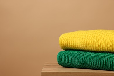 Stack of knitted sweaters on wooden table against light brown background. Space for text