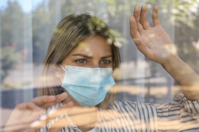 Photo of Stressed woman in protective mask looking out of window, view through glass. Self-isolation during COVID-19 pandemic