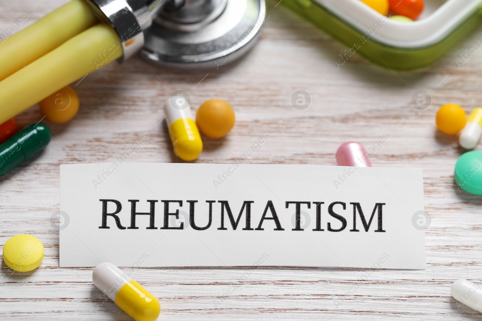 Photo of Card with word Rheumatism, stethoscope and pills on white wooden table, closeup