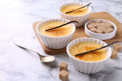 Photo of Delicious creme brulee in bowls, vanilla pods, sugar cubes and spoon on white marble table