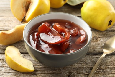 Photo of Tasty homemade quince jam in bowl, spoon and fruits on wooden table, closeup