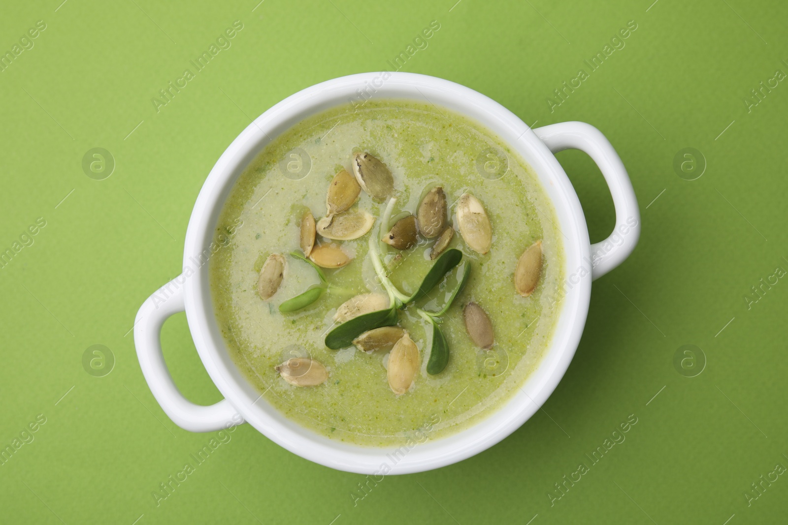 Photo of Delicious broccoli cream soup with pumpkin seeds on green background, top view