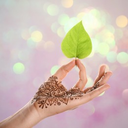 Image of Woman with henna tattoo on palm holding green leaf against blurred lights, bokeh effect