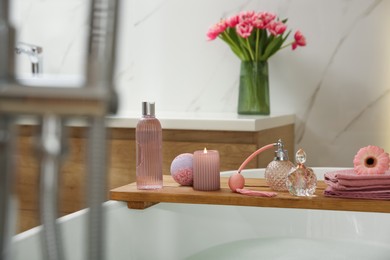 Photo of Wooden bath tray with candle and personal care products on tub indoors