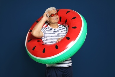Photo of Funny mature man with bright inflatable ring talking on phone on dark blue background