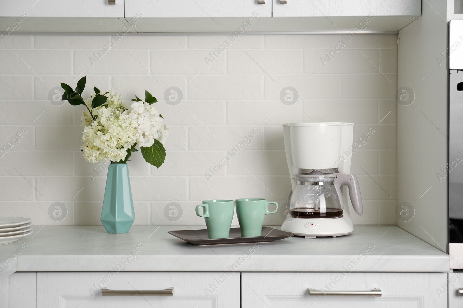 Photo of Bouquet with beautiful white hydrangea flowers, coffee maker and cups on light countertop