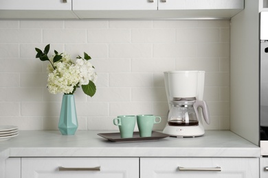 Photo of Bouquet with beautiful white hydrangea flowers, coffee maker and cups on light countertop