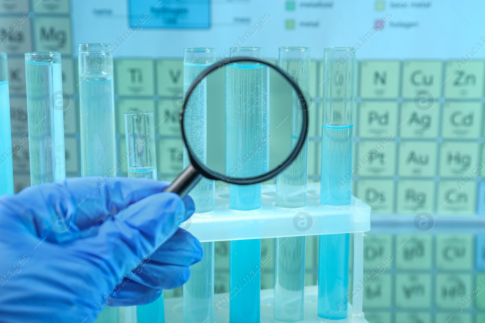 Photo of Laboratory assistant looking through magnifying glass on test tubes against periodic table of chemical elements, closeup