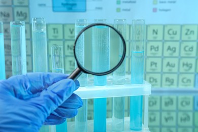 Photo of Laboratory assistant looking through magnifying glass on test tubes against periodic table of chemical elements, closeup