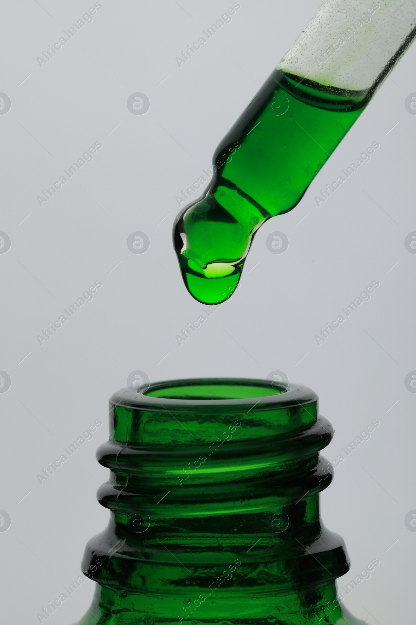 Photo of Dripping green facial serum from pipette into glass bottle on white background, closeup