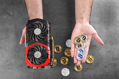Man with modern video card and bitcoins over grey table, top view