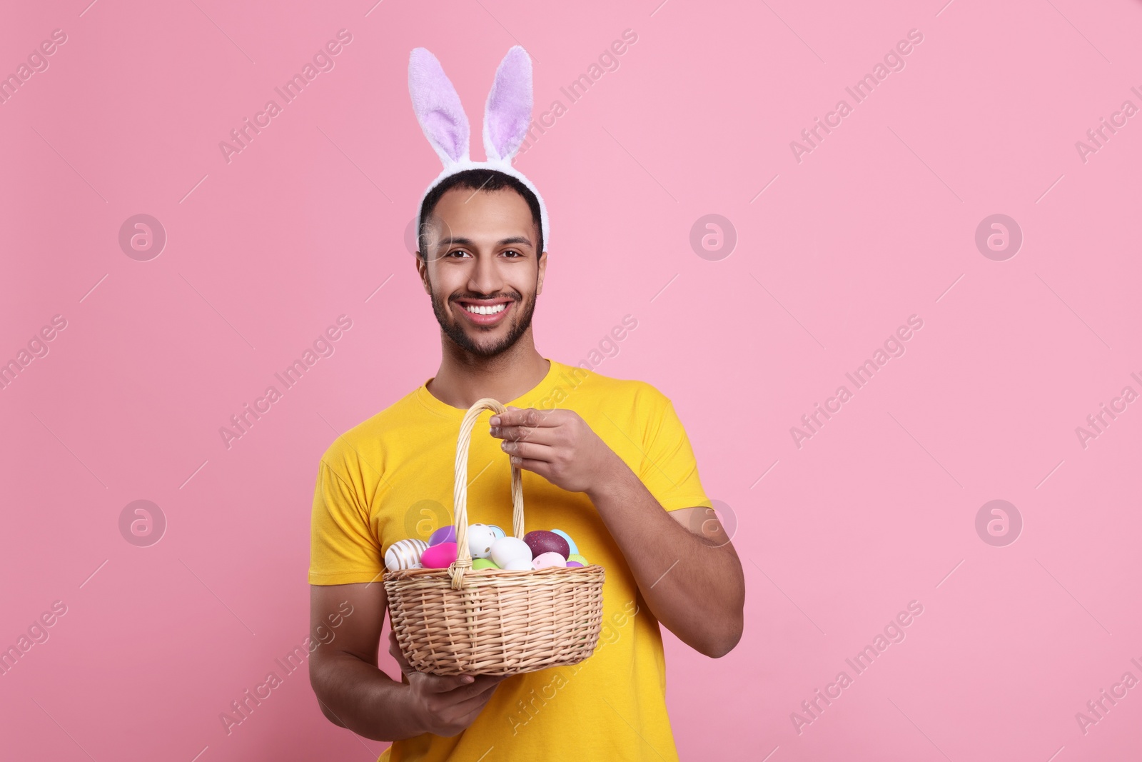 Photo of Happy African American man in bunny ears headband holding wicker basket with Easter eggs on pink background. Space for text