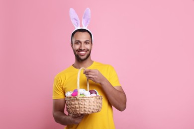 Happy African American man in bunny ears headband holding wicker basket with Easter eggs on pink background. Space for text