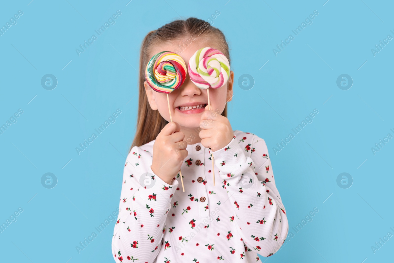 Photo of Happy little girl covering eyes with colorful lollipops on light blue background