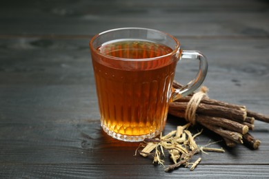 Photo of Aromatic licorice tea in cup and dried sticks of licorice root on black wooden table