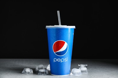 Photo of MYKOLAIV, UKRAINE - JUNE 9, 2021: Paper Pepsi cup and ice cubes on light grey table against dark background