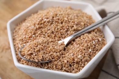 Dry wheat groats and spoon in bowl on table, closeup