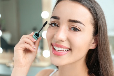 Attractive young woman applying mascara on her eyelashes against blurred background