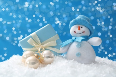 Image of Cute snowman, Christmas balls and gift on snow against blue background, bokeh effect