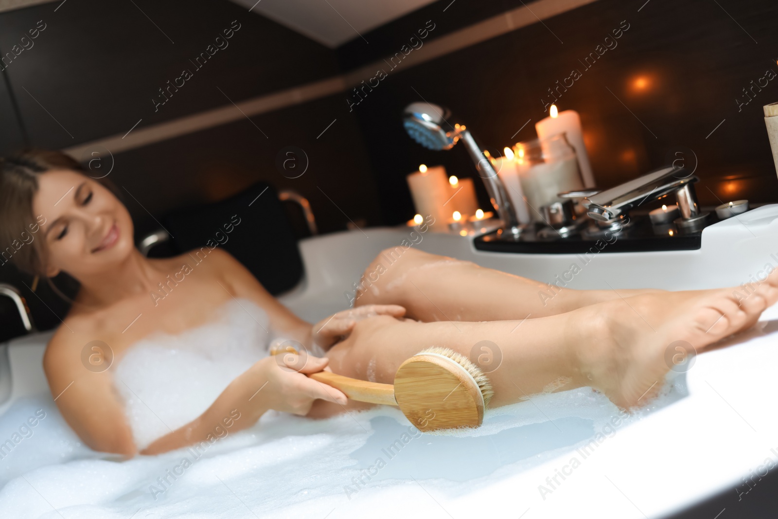 Photo of Beautiful woman rubbing leg with while taking bubble bath, focus on brush. Romantic atmosphere