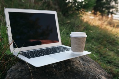 Photo of Modern laptop with blank screen and coffee cup on stone in nature, space for text. Working outdoors