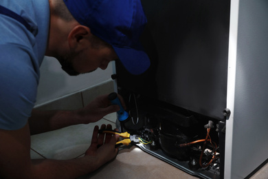 Photo of Professional electrician with flashlight fixing refrigerator indoors