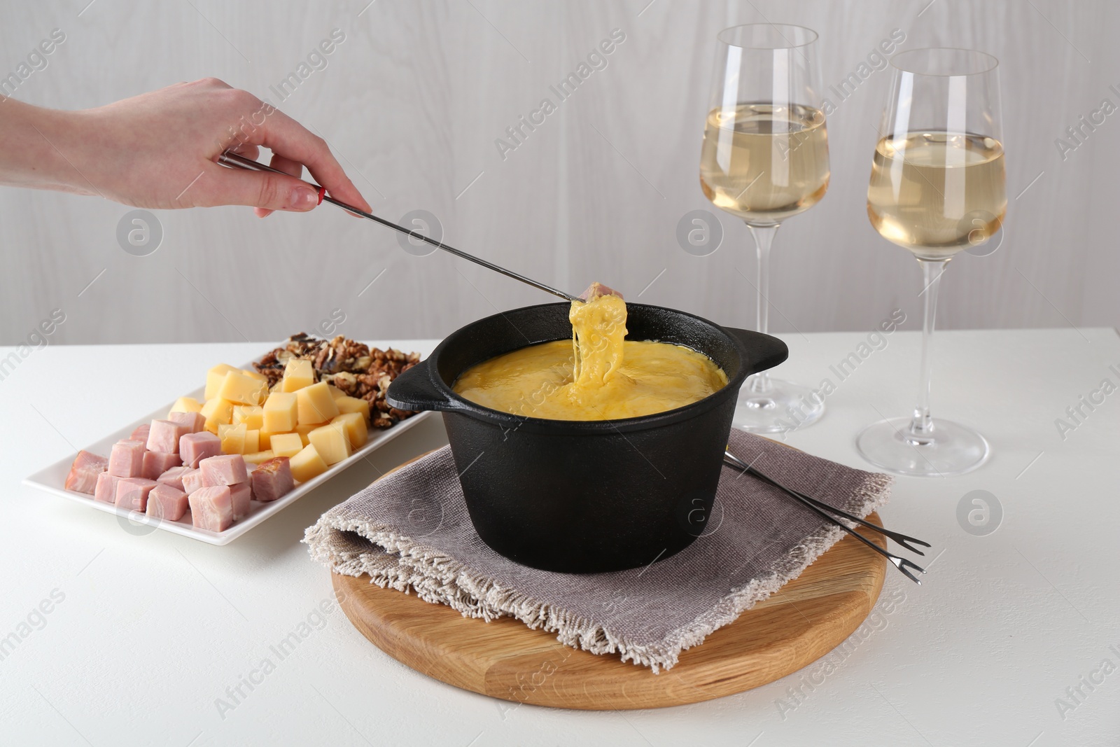 Photo of Dipping piece of ham into fondue pot with tasty melted cheese at white table, closeup