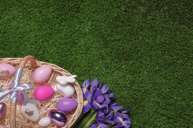 Easter basket with painted eggs, figure of rabbit and iris flowers on green grass, flat lay. Space for text