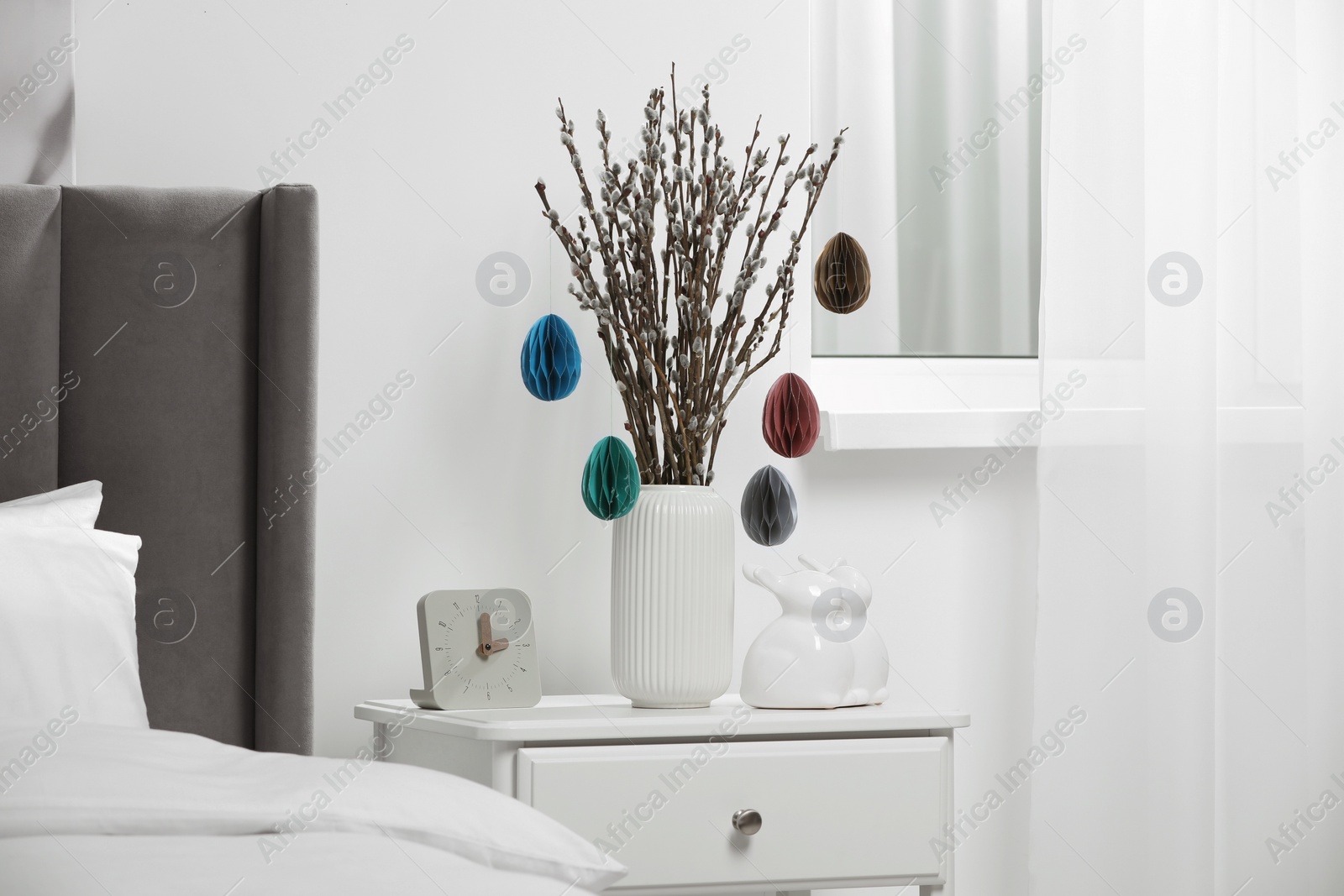 Photo of Beautiful pussy willow branches with paper eggs in vase and ceramic bunnies on nightstand at home. Easter decor