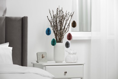 Beautiful pussy willow branches with paper eggs in vase and ceramic bunnies on nightstand at home. Easter decor