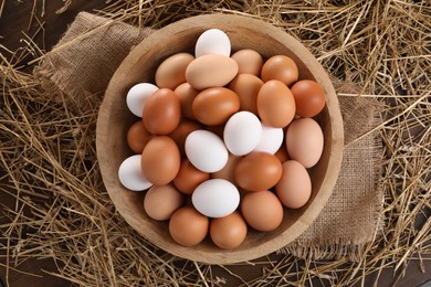 Fresh chicken eggs in bowl and dried straw on wooden table, top view