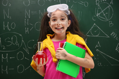 Photo of Schoolgirl holding flask and notebooks near chalkboard with chemical formulas