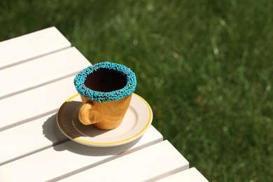 Photo of Delicious edible biscuit coffee cup decorated with sprinkles on white wooden table outdoors, space for text