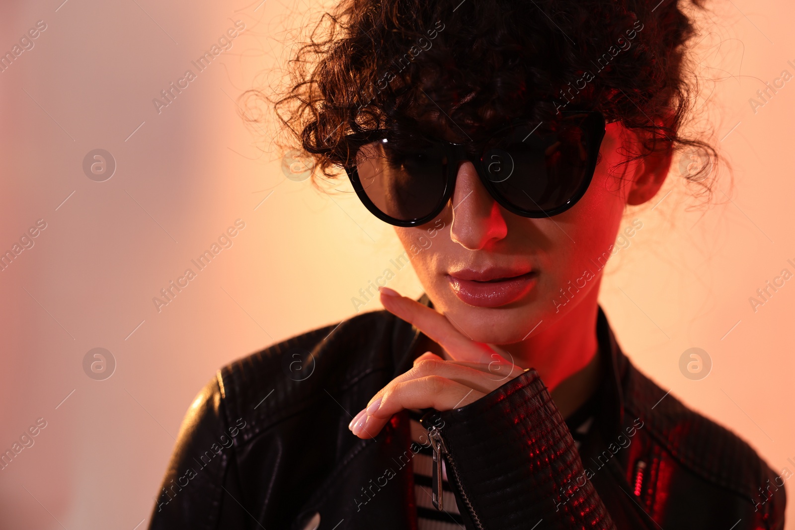 Photo of Beautiful young woman in black leather jacket and sunglasses posing on color background in neon lights, closeup