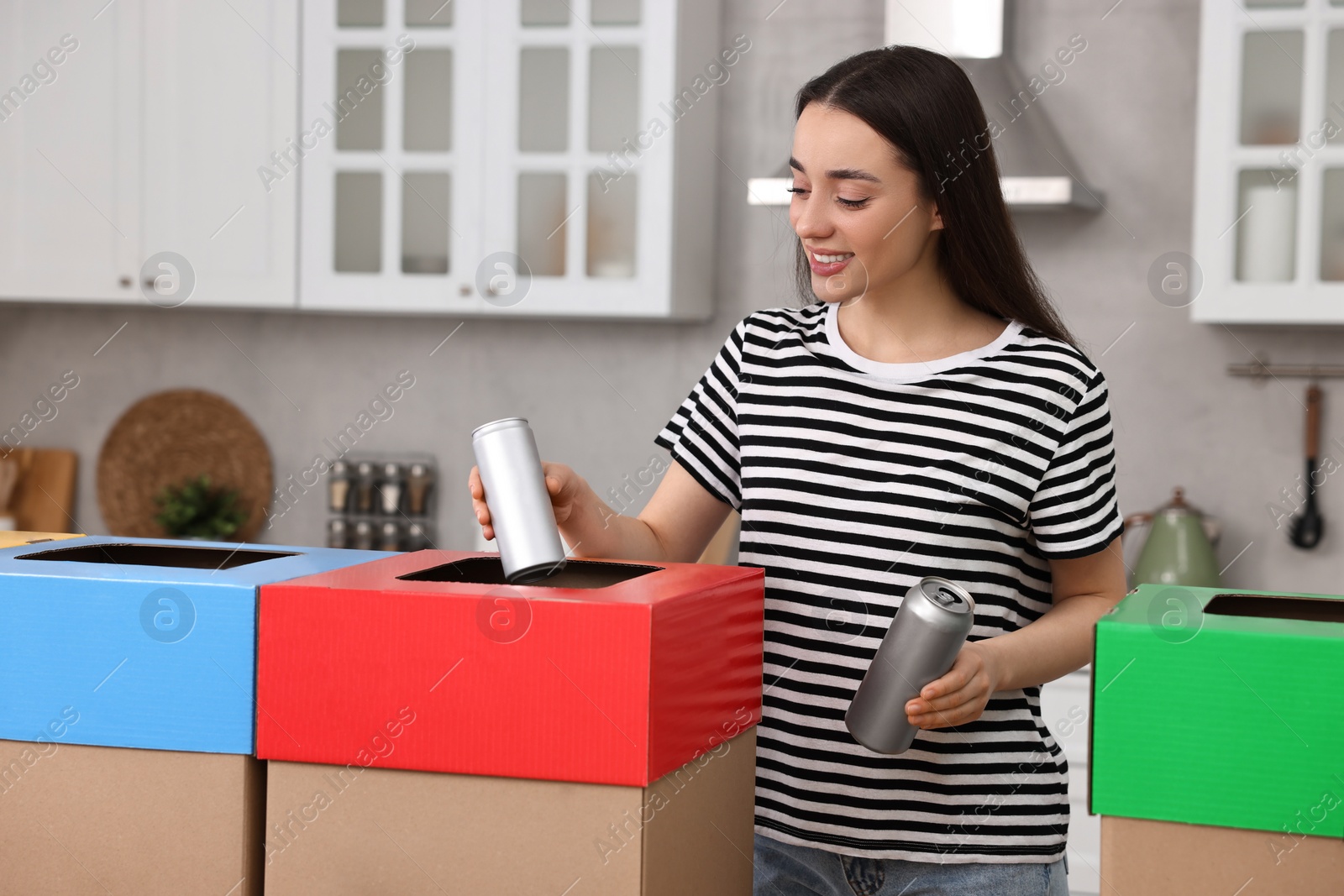 Photo of Garbage sorting. Smiling woman throwing metal can into cardboard box in kitchen