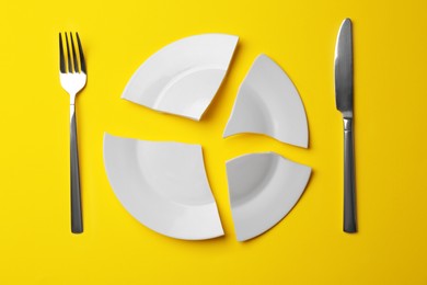 Pieces of broken white ceramic plate and cutlery on yellow background, flat lay