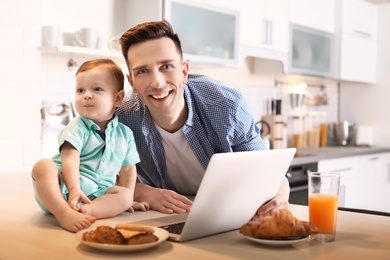 Photo of Young father with his cute little son using laptop in kitchen