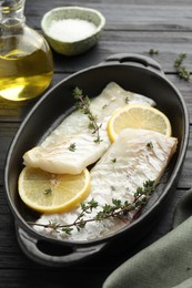 Photo of Fresh raw cod fillets with thyme and lemon in baking dish on black wooden table