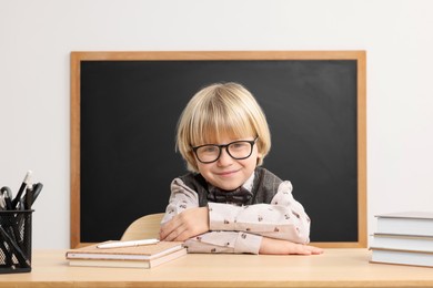 Photo of Happy little school child sitting at desk with books near chalkboard in classroom