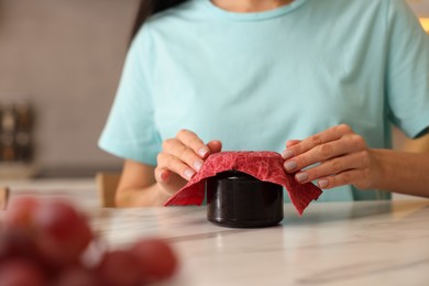 Photo of Woman packing jar of jam into beeswax food wrap at light table in kitchen, closeup