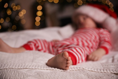 Baby in Christmas pajamas and Santa hat sleeping on bed indoors, focus on foot