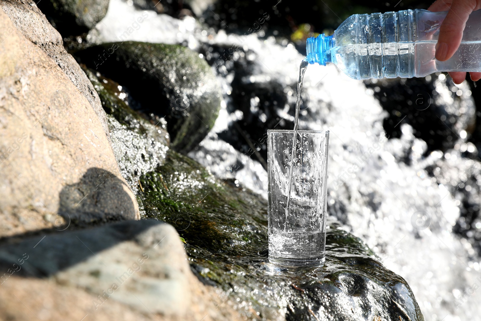 Photo of Woman pouring water from plastic bottle into glass near stream outdoors, closeup. Space for text