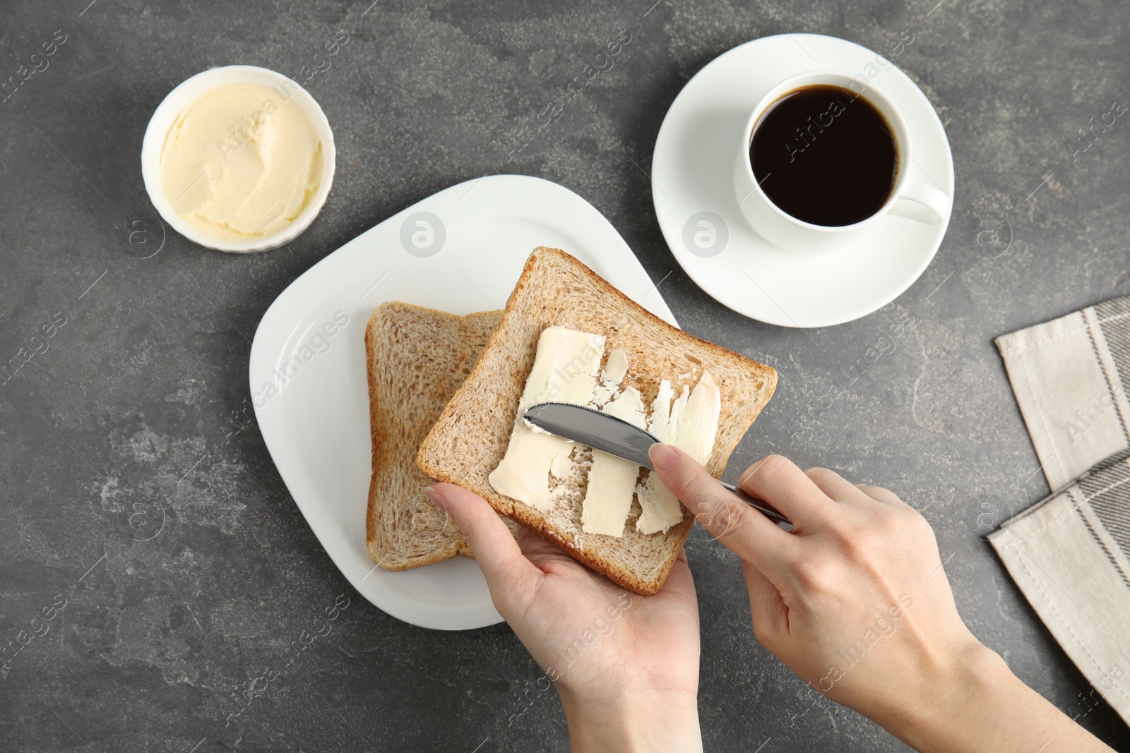 Photo of Woman spreading tasty butter onto bread over plate at grey table, top view